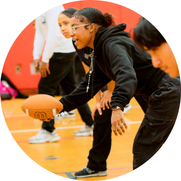 Person in black athletic wear prepares to hike a football indoors, with others standing behind on a gym floor.