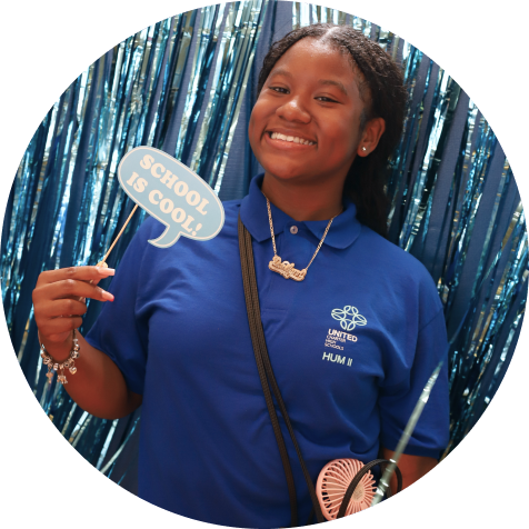 A smiling student in a blue polo shirt holds a sign that says "School is cool" in front of a blue tinsel backdrop.