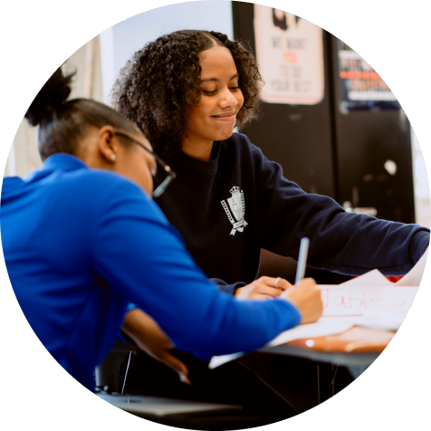Two students sit at a table working together on assignments, one writing in a notebook while the other looks on and smiles.