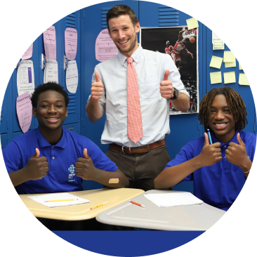 A teacher stands behind two students at desks in a classroom, all three giving thumbs up and smiling at the camera.