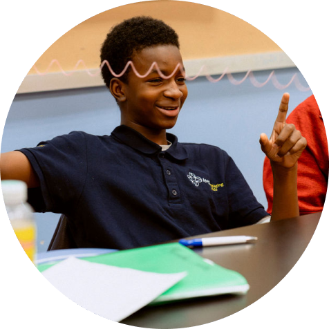 A student in a navy polo shirt sits at a desk, smiling and gesturing with one hand raised. Papers and a pen are visible on the desk in front of him.