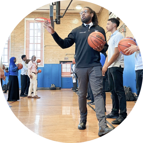 A group of people stand in a gym holding basketballs while a coach gives instructions at the front of the group.