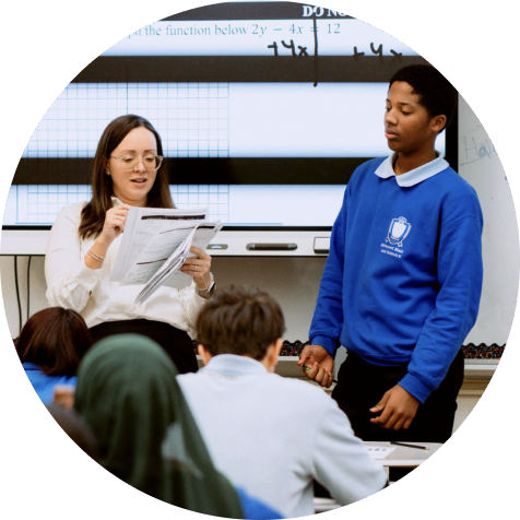 A teacher holds papers while speaking to a student in a blue uniform at the front of a classroom, with other students seated and a whiteboard in the background.