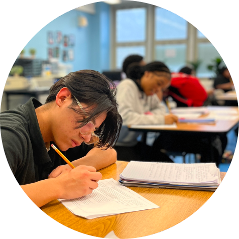 Student writing at a desk in a classroom with other students in the background also working on assignments.