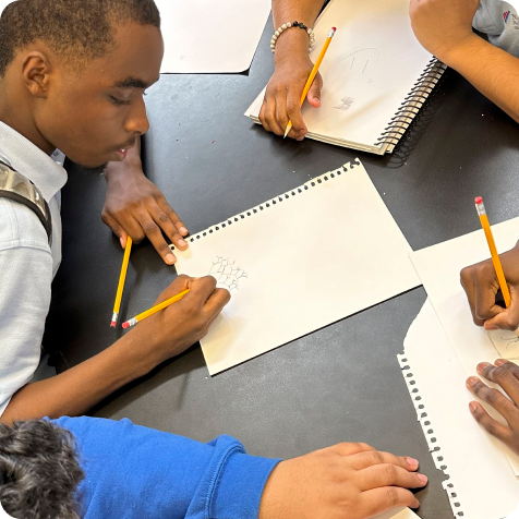 Four students seated at a table sketch on sheets of paper with pencils, focusing on their individual drawings.