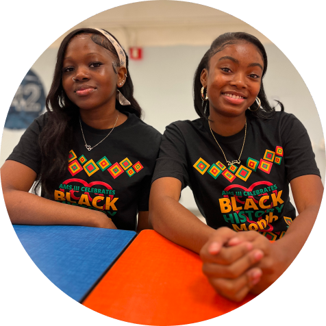 Two young women sit at a table, both wearing matching black t-shirts that celebrate Black History Month with colorful text and patterns.