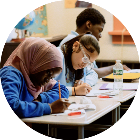 Three students sit at desks in a classroom, focused on writing assignments. School supplies and a water bottle are visible on the desks.