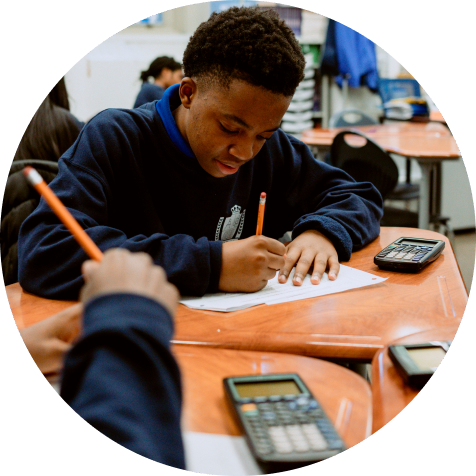 A student sits at a desk, writing on paper with a pencil. A calculator is on the desk, and other students are visible in the background.