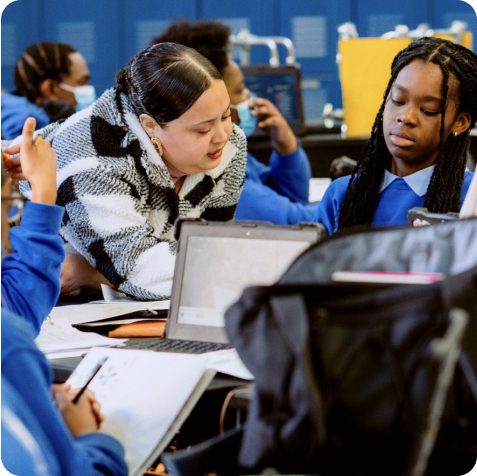 A teacher leans over to assist a student with a laptop in a classroom, while other students work at nearby desks.