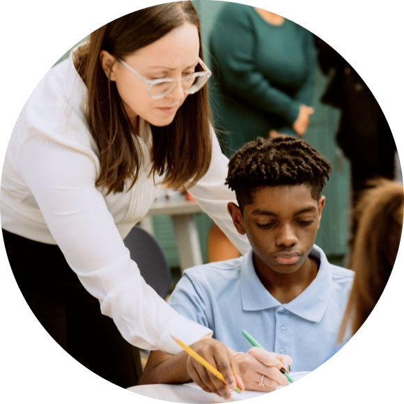 A teacher stands beside a seated student, guiding him with his work as they both focus on a worksheet at a desk in a classroom.
