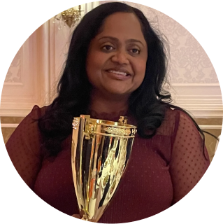 A woman with long dark hair holds a large gold trophy and smiles, standing in front of a decorative light-colored wall.