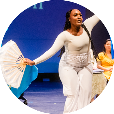 A person in a white outfit dances on stage while holding an open white fan, with another person seated in the background.