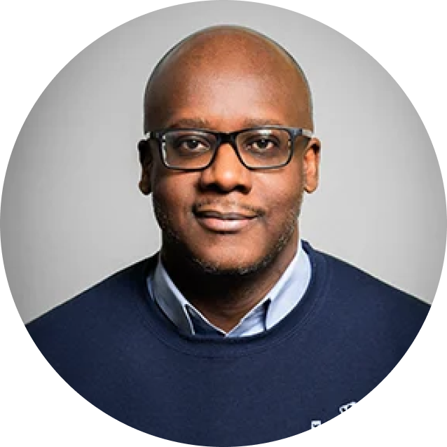 A man wearing glasses and a dark sweater over a collared shirt, facing forward and smiling slightly against a plain gray background.