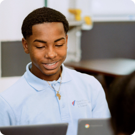 A young man in a light blue polo shirt sits at a desk, looking at a Chromebook laptop and smiling.