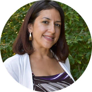 Woman with straight brown hair wearing a white cardigan and patterned top, standing outdoors in front of green foliage, smiling at the camera.