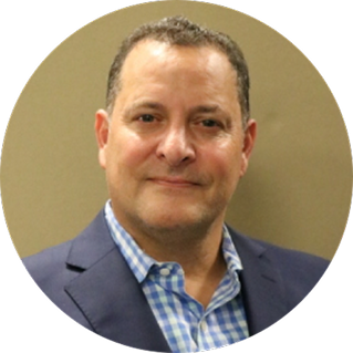 A middle-aged man wearing a navy blazer and a blue checkered shirt, posing in front of a plain beige background.