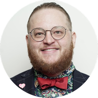 Person with glasses, short hair, and a full beard wearing a patterned shirt, red bow tie, and dark jacket with a heart-shaped pin, smiling at the camera.
