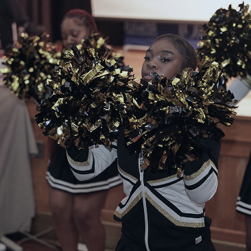 A cheerleader in a black and gold uniform holds black and gold pom-poms, performing indoors with teammates in the background.