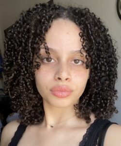 A person with shoulder-length curly hair, a gold nose ring, and a small necklace looks directly at the camera in natural light.