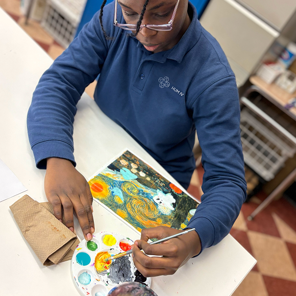 A student in a blue shirt uses a paintbrush and palette to paint a colorful picture at a table.