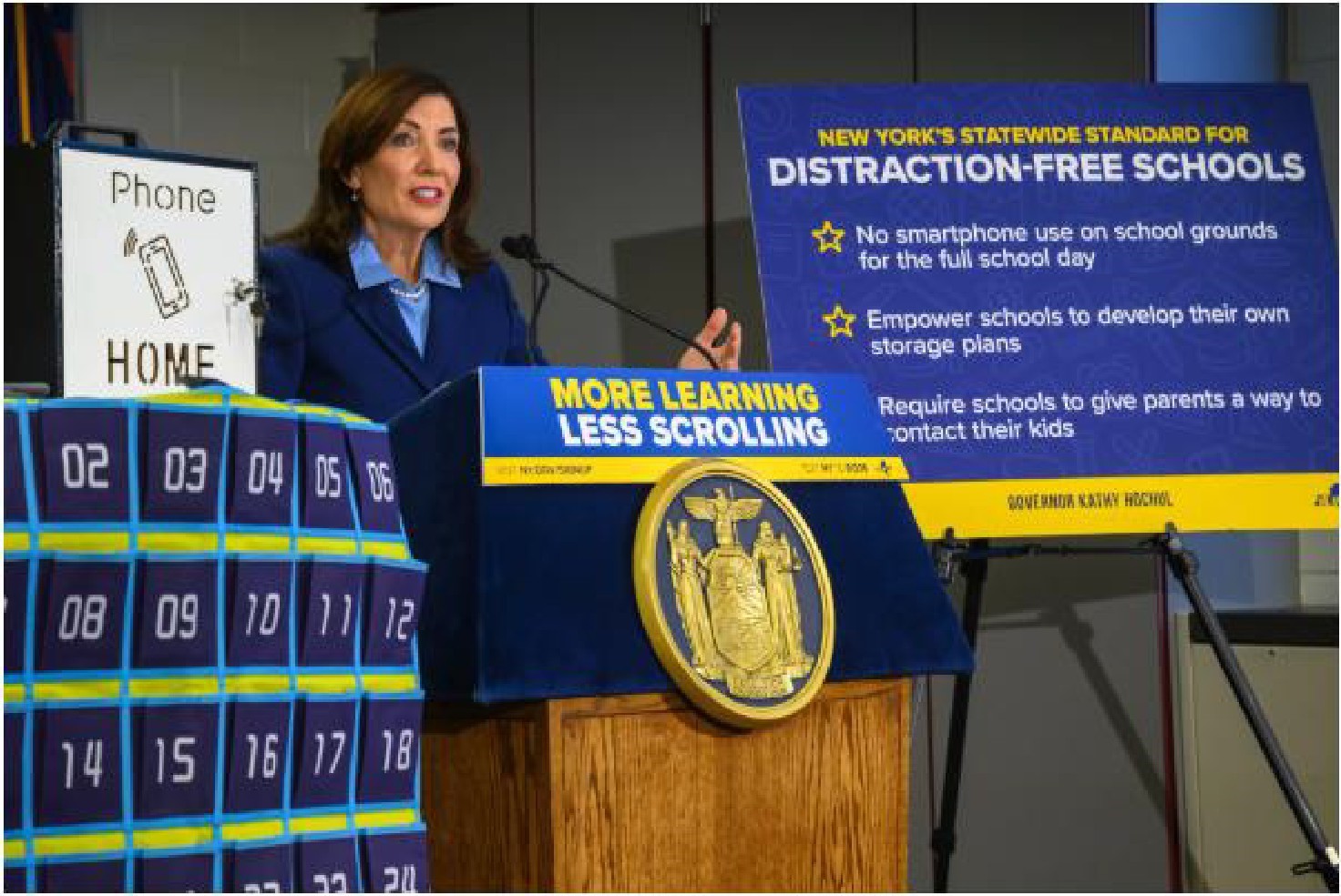A woman speaks at a podium beside a sign outlining new rules for distraction-free schools, with a phone storage station labeled “Phone Home” in the foreground.