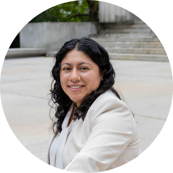 A woman with long dark curly hair wearing a white blazer sits outdoors in front of concrete steps, smiling at the camera.