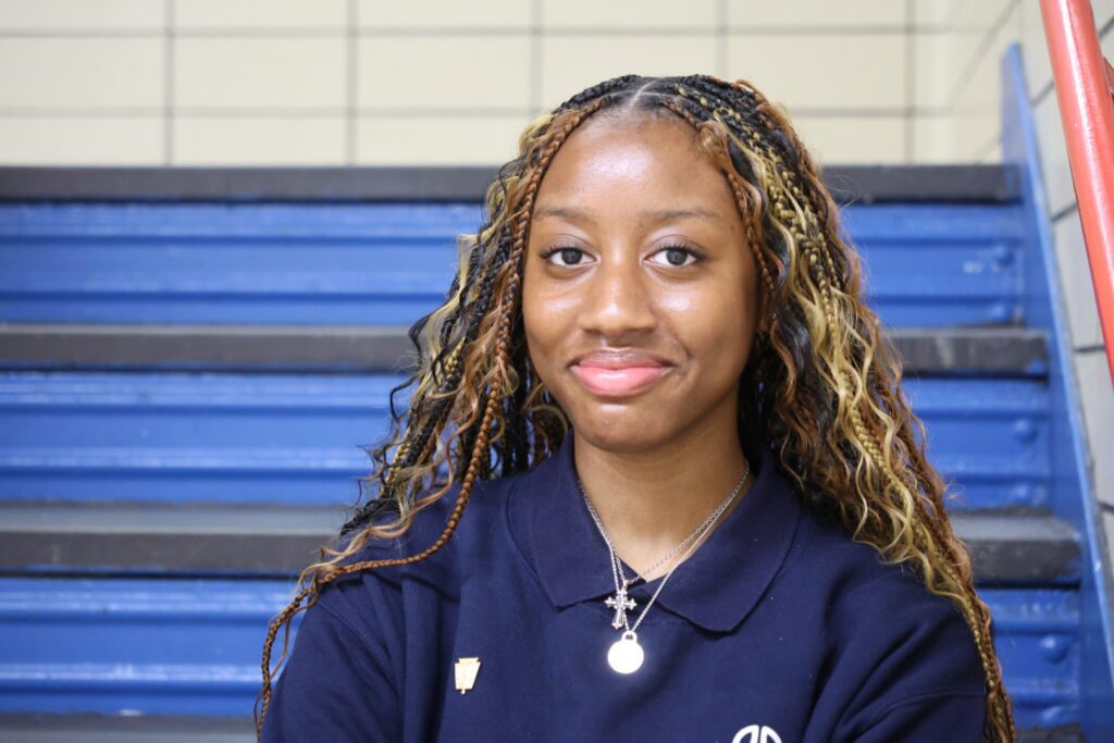 A young woman with long, braided hair and a navy polo shirt sits on blue stairs, smiling slightly at the camera.