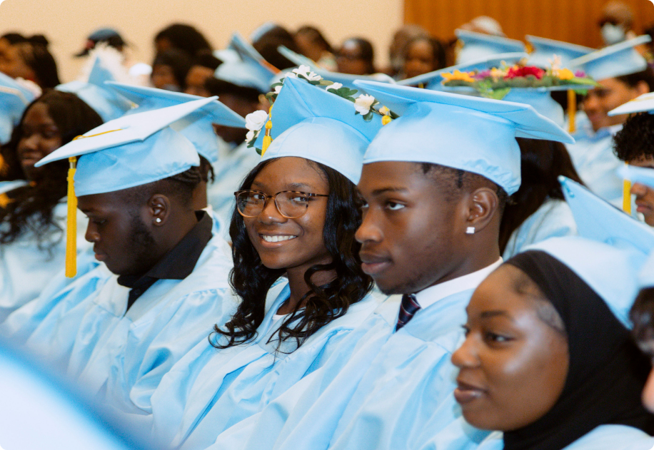 A group of students in light blue caps and gowns sit in rows at a graduation ceremony, some smiling and some looking ahead.