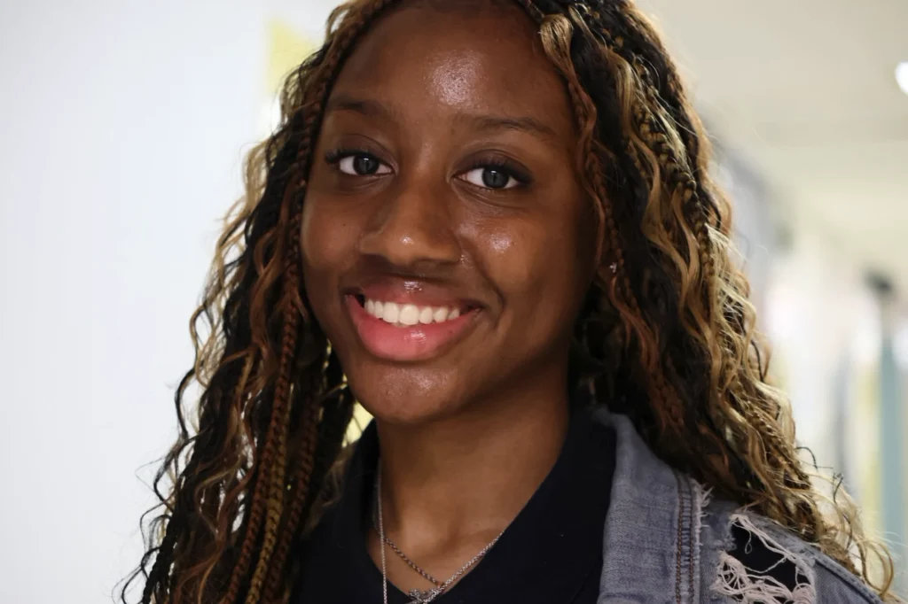 A young woman with long, wavy hair and a nose ring smiles at the camera indoors, wearing a denim jacket over a dark shirt.