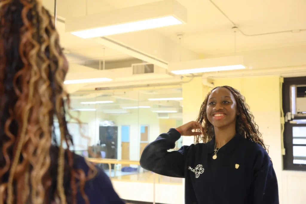 A woman with braided hair stands in front of a mirror, smiling and adjusting her hair in a brightly lit room with yellow walls.