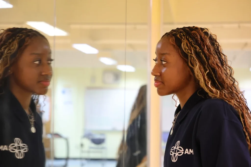 A woman with braided hair stands indoors, looking at her reflection in a large mirror.