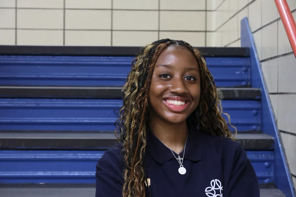 A young woman with long braids and a navy shirt smiles while sitting on blue stairs in a tiled indoor setting.