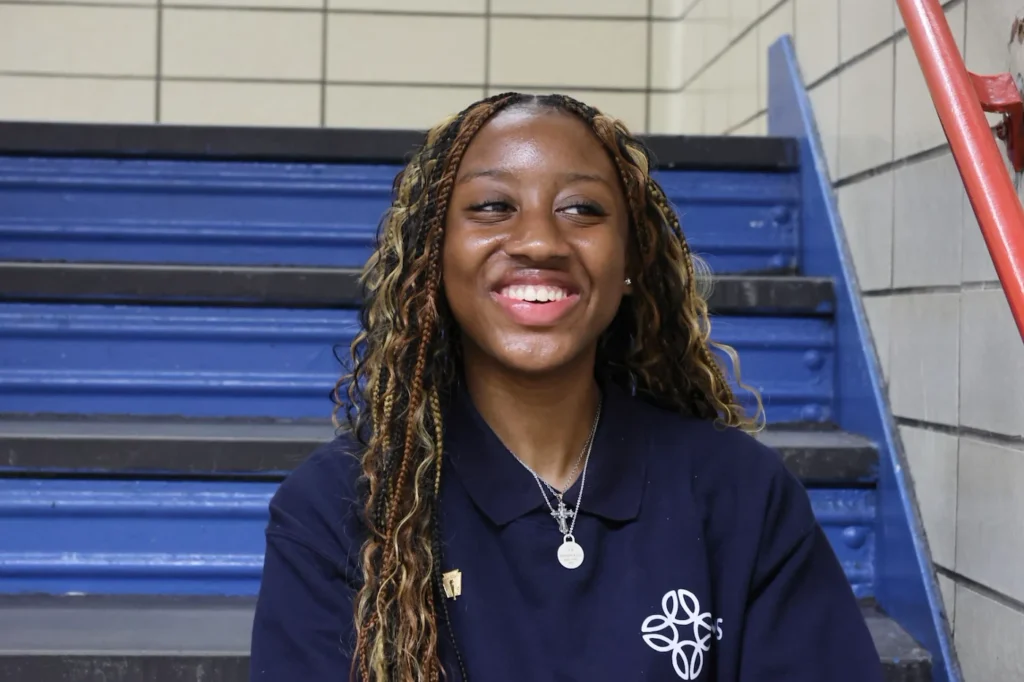 A young woman with long braids, wearing a navy blue collared shirt, sits and smiles on blue stairs in a tiled indoor setting.