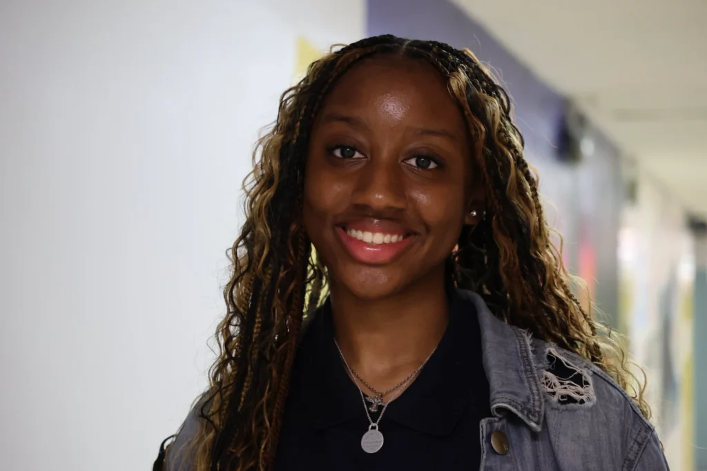 A young woman with long, curly hair and a necklace smiles at the camera while standing in a hallway with blurred walls in the background.