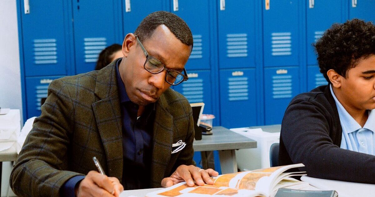 An adult man in glasses is writing in a book at a desk next to a student in a classroom with blue lockers in the background.
