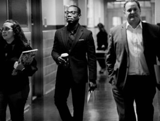 Three professionally dressed people walk down a hallway; one holds a coffee cup, another carries a notebook, and the third has a folder. The image is in black and white.