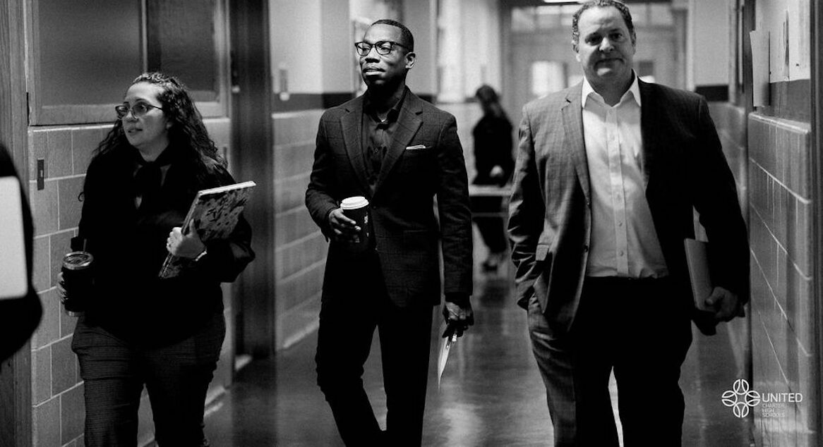 Three professionally dressed people walk down a hallway; one holds a coffee cup, another carries a notebook, and the third has a folder. The image is in black and white.