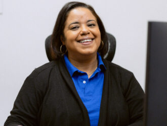A United Charter High Schools in a blue shirt and black cardigan sits at a desk, smiling at the camera with one hand on a computer mouse.