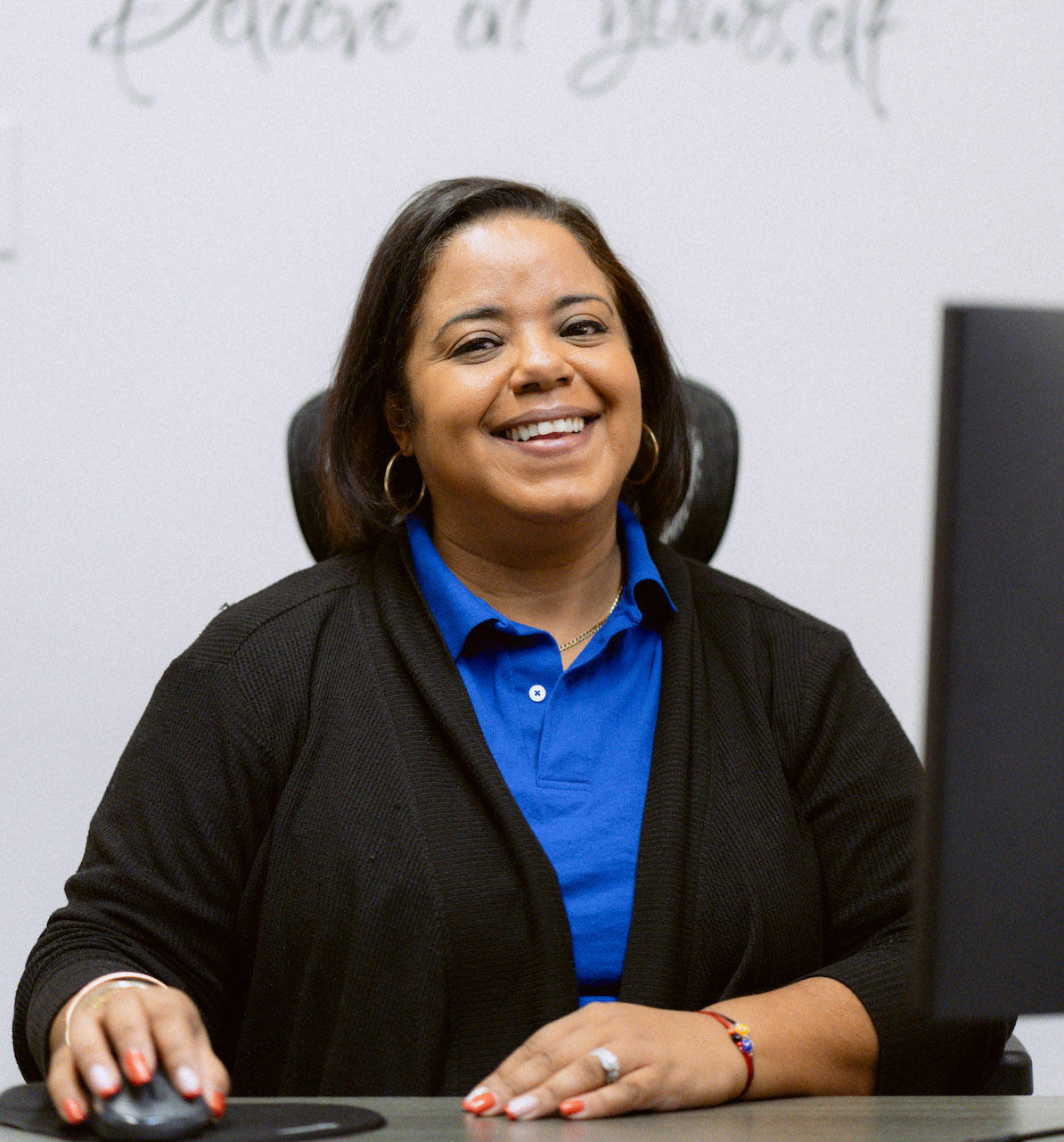 A United Charter High Schools in a blue shirt and black cardigan sits at a desk, smiling at the camera with one hand on a computer mouse.