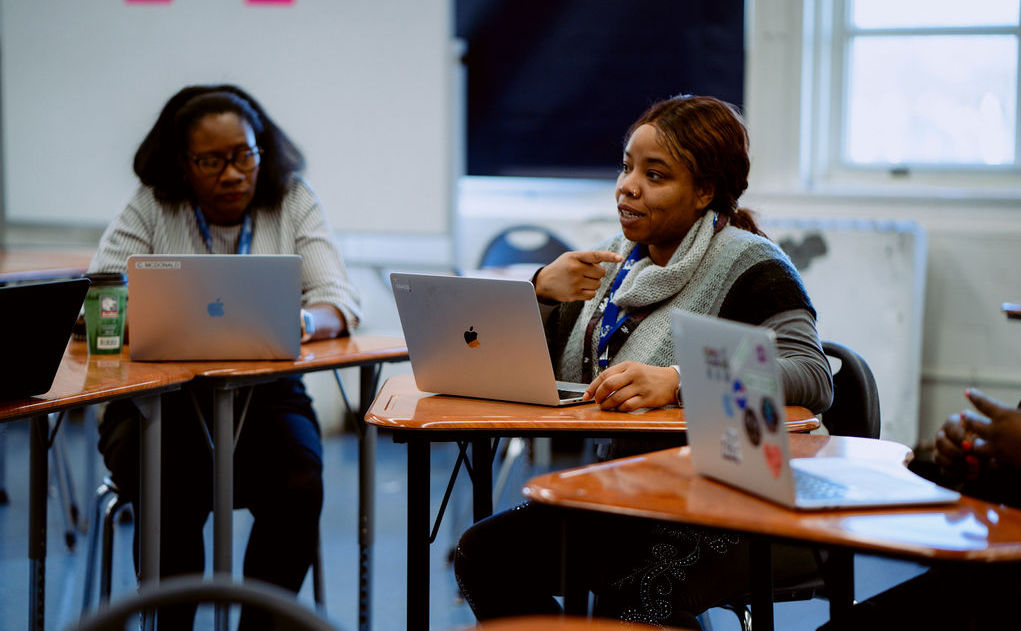 wo staff members at United Charter High Schools sit at classroom desks with laptop computers.