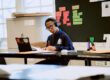 A United Charter High School student wearing headphones sits at a desk, writing on paper while looking at a tablet in a classroom with colorful notes on the board.