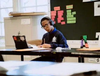 A United Charter High School student wearing headphones sits at a desk, writing on paper while looking at a tablet in a classroom with colorful notes on the board.