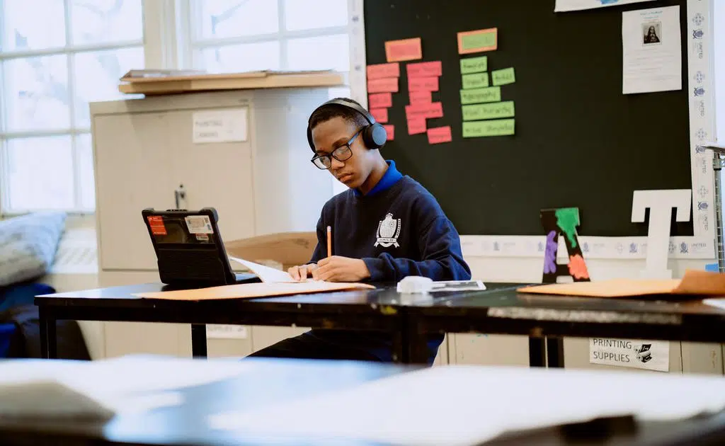 A United Charter High School student wearing headphones sits at a desk, writing on paper while looking at a tablet in a classroom with colorful notes on the board.