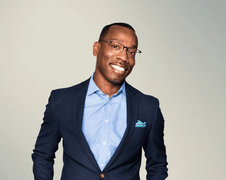 A man in a navy suit and light blue shirt stands smiling against a neutral gradient background. He wears glasses and has a pocket square in his suit jacket.