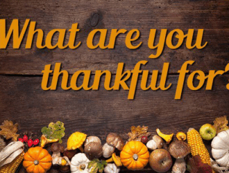 Text "What are you thankful for?" on a wooden background, with assorted autumn decorations including pumpkins, corn, and leaves arranged along the bottom.