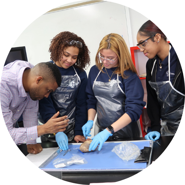 A teacher instructs three students wearing gloves and safety glasses as they dissect a specimen on a tray in a science classroom.