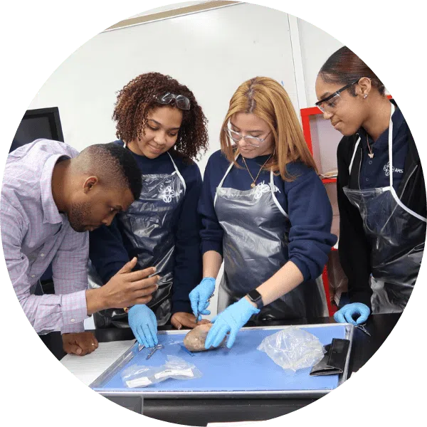 A teacher instructs three students wearing gloves and safety glasses as they dissect a specimen on a tray in a science classroom.