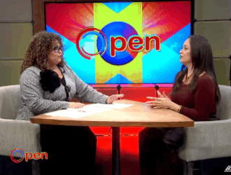 Two women sit at a round table having a discussion on a brightly lit TV set with a screen displaying the word "Open" in the background.