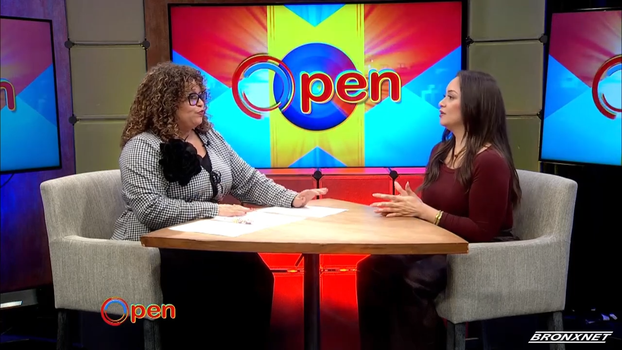 Two women sit at a round table having a discussion on a brightly lit TV set with a screen displaying the word "Open" in the background.