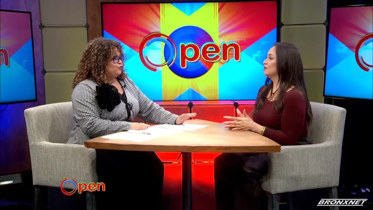 Two women sit at a round table having a discussion on a brightly lit TV set with a screen displaying the word "Open" in the background.
